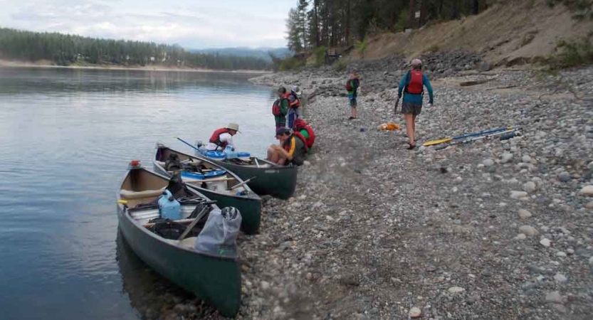 a few kayaks rest on the shore of a lake while people carry gear away from them 